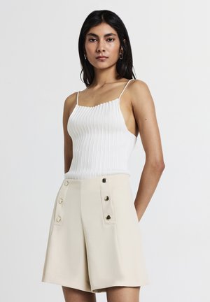 Frau mit langen, dunklen Haaren, die ein weißes, geripptes, ärmelloses Oberteil und beige, hoch taillierte Shorts mit Knopfdetails trägt, steht vor einem weißen Hintergrund.