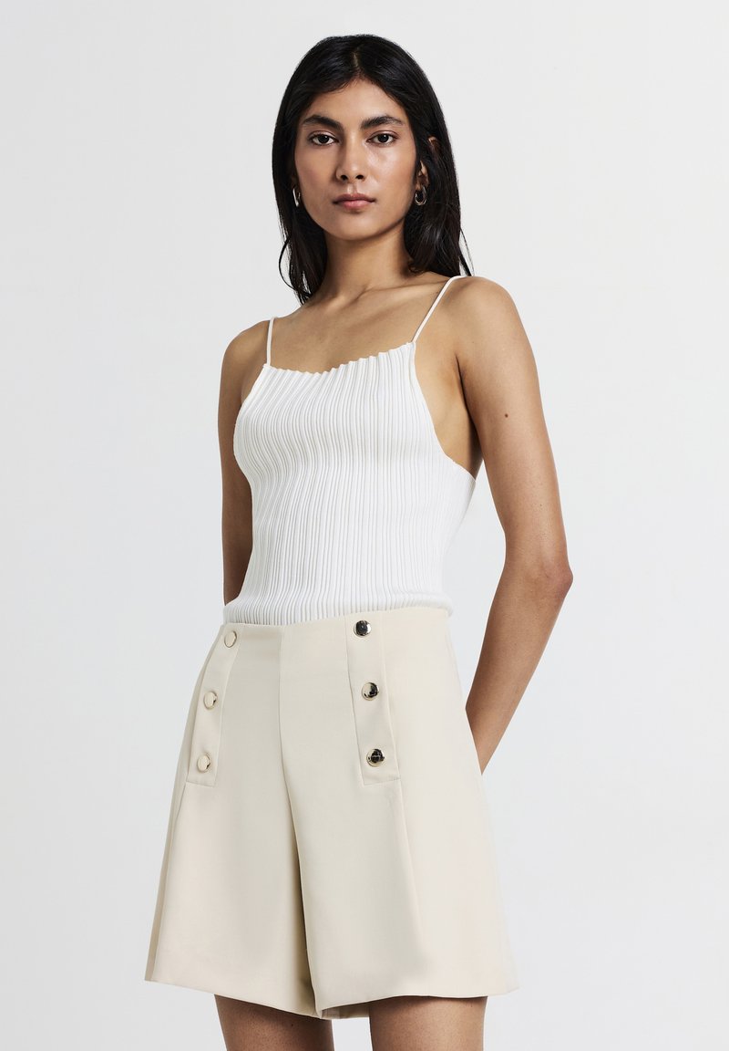 Femme avec de longs cheveux noirs portant un haut à manches courtes côtelé blanc et un short beige taille haute avec des boutons, se tenant devant un fond blanc.