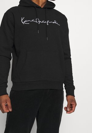 Homme portant un sweat à capuche noir avec un texte en cursive blanc et un pantalon en velours côtelé noir, debout devant un fond clair uni.