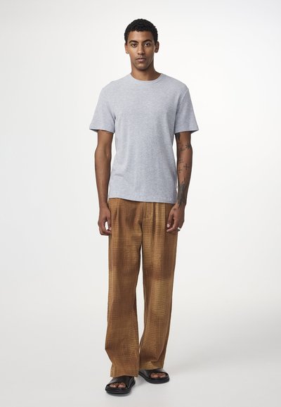 Grå bomulds T-shirt med rund hals, parret med brune mønstrede bukser med vidde og sorte sandaler, stående op mod en neutral baggrund.