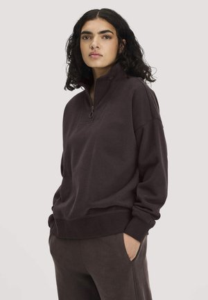 Vrouw met krullend donker haar, die een donkerbruine trui met kwartritssluiting en bijpassende broek draagt, handen in de zakken op een lichte achtergrond.