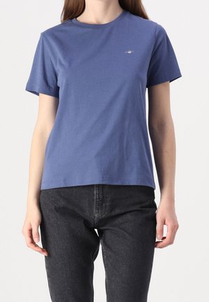 Femme portant un t-shirt bleu uni à manches courtes avec un petit logo sur la poitrine et un jean noir, debout devant un fond uni.