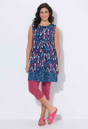 Mujer sonriente de pie con las piernas cruzadas, vestida con un túnico sin mangas de patrón azul, leggings cortos rosas y sandalias rosas con hebilla.