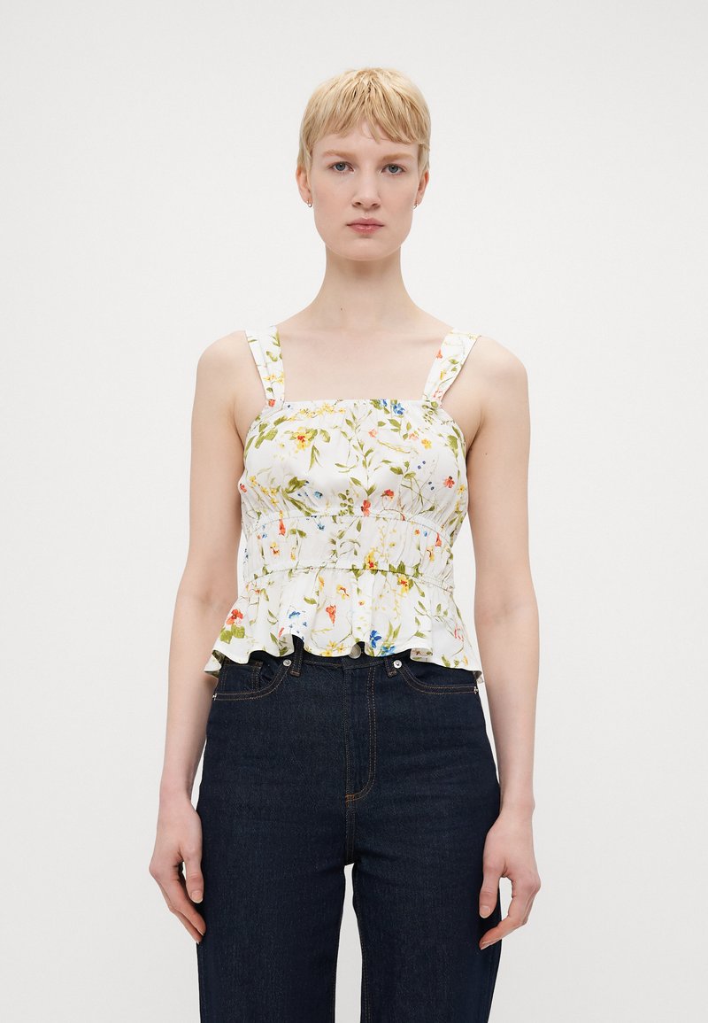 Blomstrete peplum-topp med hvit bakgrunn, med fargerike blomstermønstre, brede stropper og volangkant. Kombinert med mørke denimjeans.