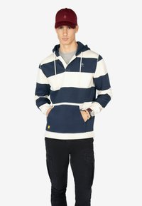 Sudadera a rayas en navy y crema con capucha ajustable, bolsillo frontal y detalle de logo. Combinada con pantalones cargo negros y una gorra color burdeos.