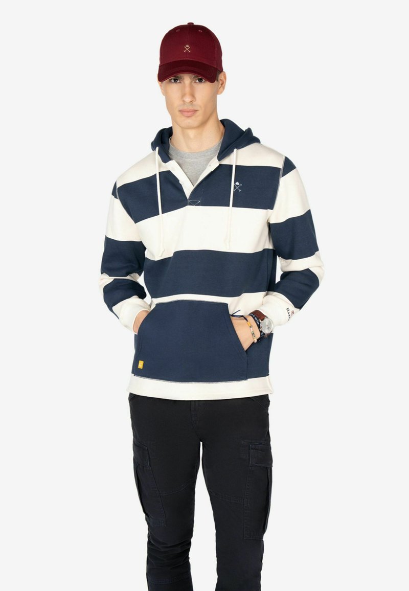 Sudadera a rayas en navy y crema con capucha ajustable, bolsillo frontal y detalle de logo. Combinada con pantalones cargo negros y una gorra color burdeos.