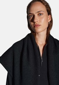 Coprispalle nero in maglia con una texture morbida e un leggero scintillio, drappeggiato su una maglia nera con cerniera. Il design presenta una forma ampia e fluida.