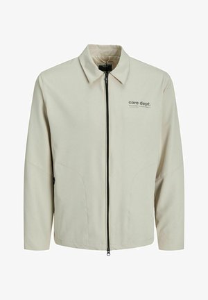 Beige színű, cipzáras dzseki hosszú ujjal, hegyes gallérral és két oldalsó zsebbel. Bal mellen logó található. Simaság jellemzi az anyag textúráját.