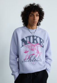 Világoslila pulóver, amelyen egy szárnyas ló grafika és a "NIKE ATHLETICS" felirat található fekete és rózsaszín színben. Puha anyagú, bordás mandzsettákkal.