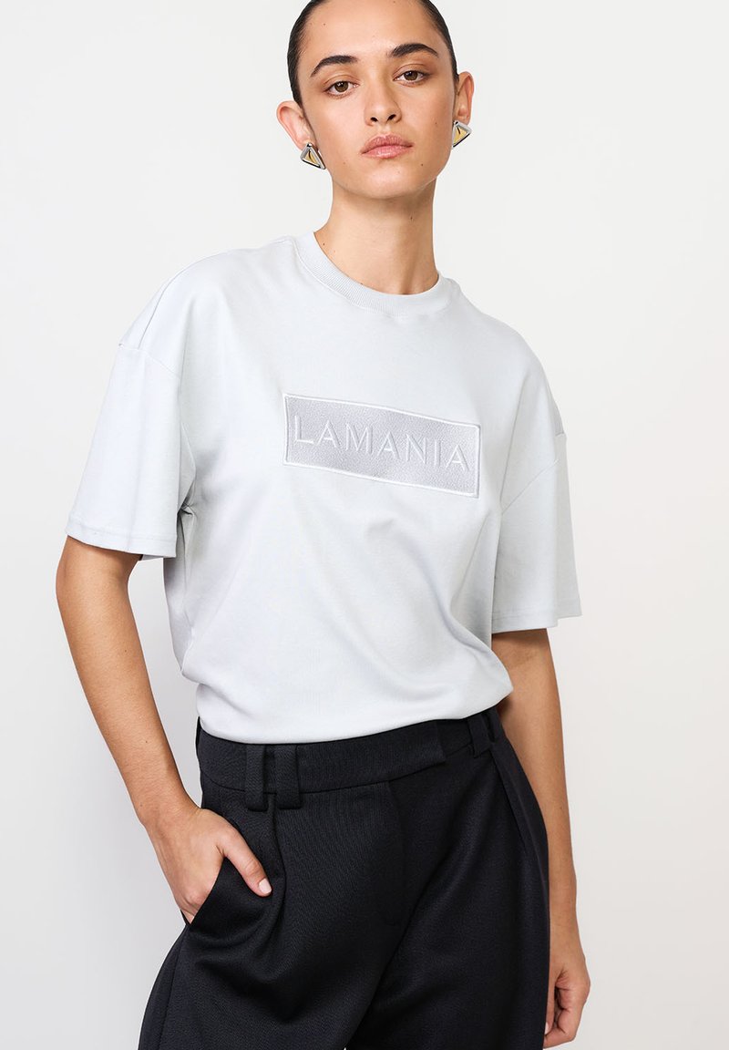 Šviesiai pilkas medvilninis marškinėlis su iškeltu 'LAMANIA' logotipu, laisvas kirpimas, trumpomis rankovėmis, derinamas su juodomis aukštu liemeniu kelnėmis.