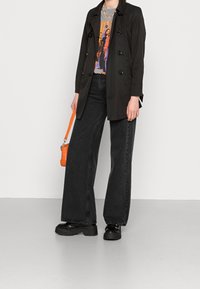 Svart trenchcoat med dubbelknappning, tillsammans med en grafisk t-shirt. Vidbeniga svarta jeans och kraftiga svarta platåskor, bärande på en orange väska.