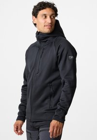 Man wearing black zip-up hooded jacket with side and chest zip pockets, looking to his left against a plain light background.