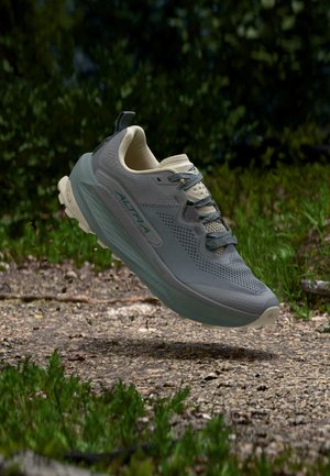 Hellgrauer Altra Laufschuh mit strukturierter Sohle, der über einem Erdweg schwebt, umgeben von grünem Gras und Pflanzen.