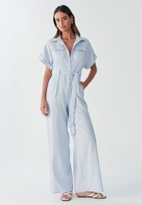 Mono de botones azul claro con mangas cortas, dos bolsillos en el pecho, cintura con cinturón y pantalones de pierna ancha. Tejido liso y ligero.