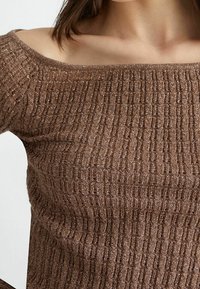 Ruskea teksturoitu olkapäiltä avonainen neulepaita, jota käyttää henkilö, näyttää ylävartalon ja kaula-aukkoa.