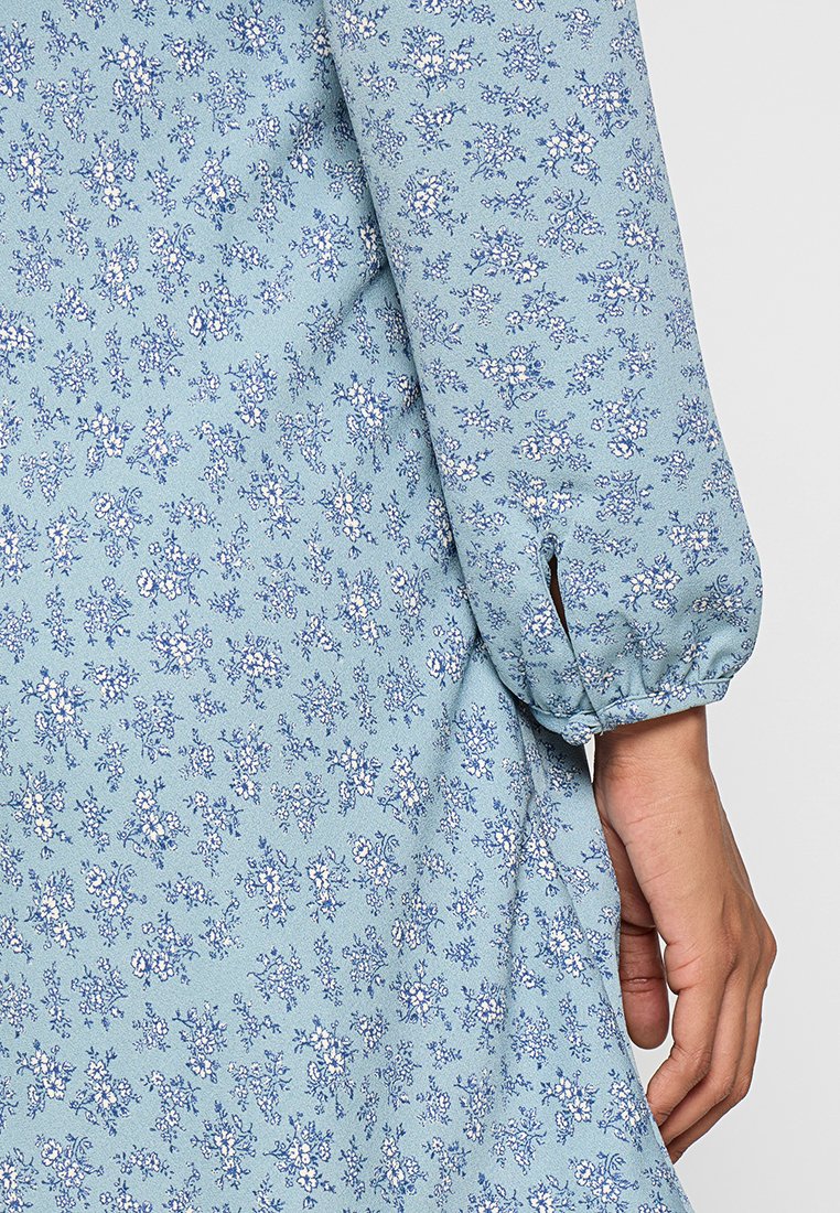 Vestido de tela con estampado floral azul y mangas largas abullonadas. El patrón presenta pequeñas flores blancas y azul oscuro sobre un fondo azul claro.