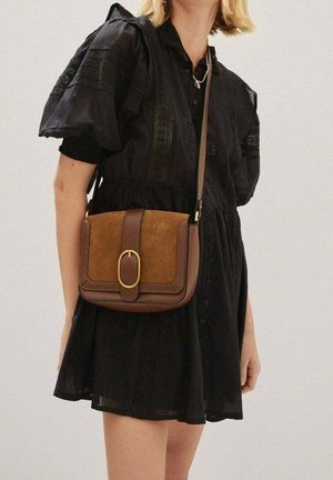 Kvinde i en sort kortærmet kjole med en brun crossbody taske med en guldfarvet oval spænde foran.