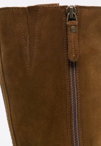 Section de botte en daim marron montrant un détail en V cousu et une fermeture éclair en métal avec une languette en daim sur fond clair.