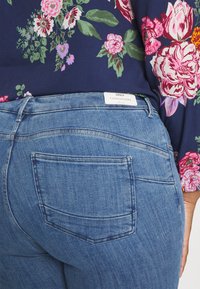Blusa escura com padrão floral, combinada com jeans de denim azul claro, com costuras visíveis e bolsos traseiros. Etiqueta com a inscrição "ONLY CARMAKOMA" na cintura.