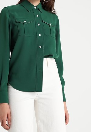 Femme portant une chemise boutonnée à manches longues vert foncé avec poches poitrine et un pantalon blanc taille haute, sur un fond uni.