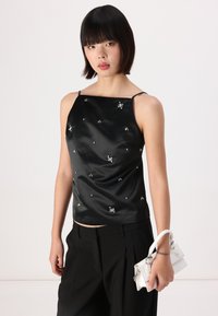 Black satin tank top with thin straps, featuring silver embellishments. Paired with high-waisted black trousers and a small white handbag.