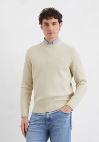 Beige gerippter Pullover mit Rundhalsausschnitt, langen Ärmeln und einem subtilen Texturmuster. Getragen über einem hellblauen gestreiften Hemd mit Kragen.