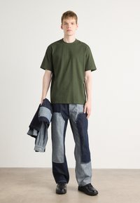 Grünes Baumwoll-T-Shirt, entspannte Passform. Blaue Jeanshose in verschiedenen Tönen mit Patchwork-Design. Modell hält eine blaue Jeansjacke und trägt schwarze Schuhe.