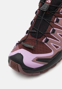 Tekaška obutev za trail running z bordo mrežastim zgornjim delom, rožnatimi akcenti, črnim podplatom in vgrajenim sistemom vezalk za varen oprijem.