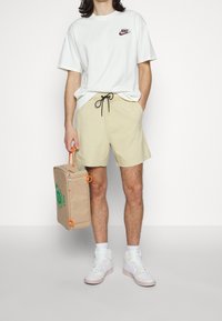 Vit Nike-t-shirt med röd logotyp, beiga shorts, vita sneakers med rosa detaljer och en beige väska med en grön spiraldesign och orangea detaljer.