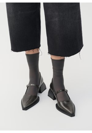 Czarne skórzane buty Mary Jane z wąskim noskiem, słupkiem i regulowanym paskiem. Noszone z przezroczystymi czarnymi skarpetkami i przetartymi dżinsami.