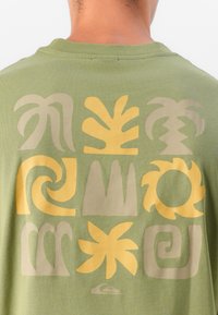 Olīvzaļš T-krekls, kam uz muguras ir izteikti dzelteni un gaiši bēši abstrakti raksti, tostarp spirāles un lapu formas.