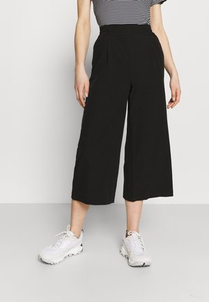 Person trägt schwarze Culotte-Hosen mit weitem Bein, ein kurzärmeliges, gestreiftes Oberteil und weiße Sneakers und steht vor einem schlichten Hintergrund.
