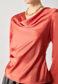 Blusa di raso corallo con scollatura drappeggiata e maniche lunghe, caratterizzata da una texture liscia e una vestibilità morbida, abbinata a una collana in oro.