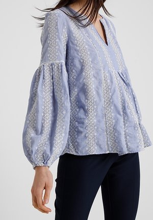 Vrouw die een lichtblauwe blouse met witte bloemenborduursel en volumineuze lange mouwen draagt, gecombineerd met een donkere broek.