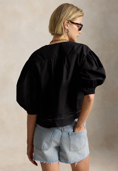 Γυναίκα με κοντά ξανθά μαλλιά που φοράει γυαλιά ηλίου καφέ τύπου χελώνας, κρίκους σκουλαρίκια, πολύχρωμο κολιέ με χάντρες, μαύρο τοπ με φουσκωτά μανίκια και ανοιχτόχρωμο τζιν σορτς.