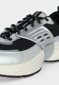 Μαύρο και ασημί αθλητικό παπούτσι Jordan με υφασμένο διχτυωτό ύφασμα, παχιά λευκή σόλα, μαύρα κορδόνια με λευκές λεπτομέρειες και μεταλλικό πλαϊνό πάνελ με διακοσμήσεις.