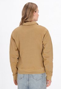 Barna kordbársony kabát, gallérral és hosszú ujjakkal, enyhén texturált felülettel és bordás mandzsettákkal, világoskék farmernadrággal párosítva.