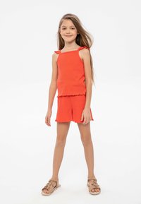 Rotes geripptes Tanktop mit Rüschen-Trägern, kombiniert mit passenden Shorts mit Rüschensaum, abgerundet mit hellbraunen Sandalen.