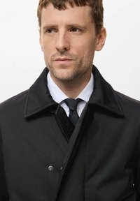 Abrigo negro hecho de una tela suave con un cuello, con cierre de botones y llevado sobre una camisa blanca y una corbata negra.