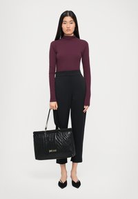 Black textured leather tote bag with gold logo, held by a person in a fitted burgundy long-sleeve top and tailored black pants.