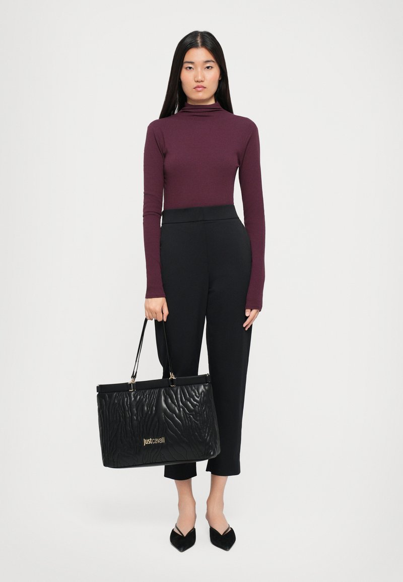 Black textured leather tote bag with gold logo, held by a person in a fitted burgundy long-sleeve top and tailored black pants.