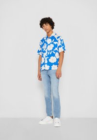 Korte mouwen blauwe bloemenhemd met grote witte bloemen, gecombineerd met lichtblauwe jeans en witte sneakers, staand tegen een neutrale achtergrond.