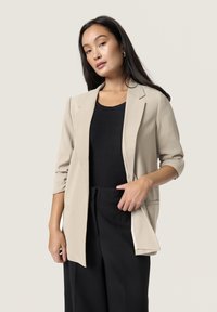 Blazer beige con un design strutturato, tasca frontale singola e maniche arrotolate, indossato sopra un top nero a costine e pantaloni neri.