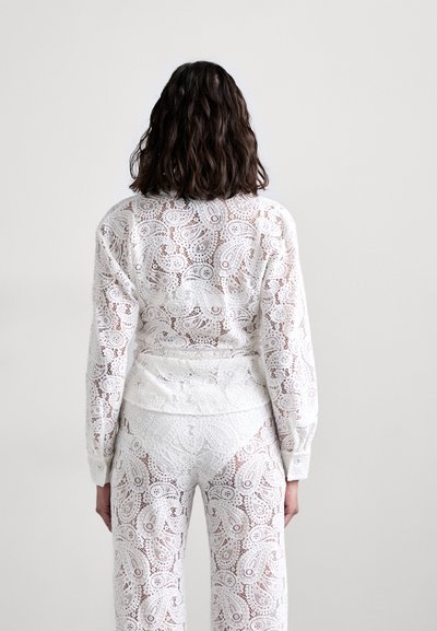 Ensemble en dentelle blanche composé d'un haut à manches longues et d'un pantalon, avec des motifs paisley et floraux intricats. Le tissu est transparent avec un détail texturé.