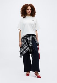 Νεαρή γυναίκα με σγουρά μαλλιά, φοράει λευκό φαρδύ t-shirt, μαύρο παντελόνι με φαρδιές σκέλη, καρό πουκάμισο δεμένο γύρω από τη μέση, κόκκινα τακούνια και κρατά κόκκινη τσάντα.