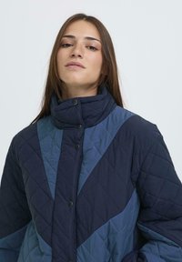 Chaqueta acolchada con un patrón diagonal en azul marino oscuro y azul claro, con un cuello alto y cierre de botones a presión. Textura suave.