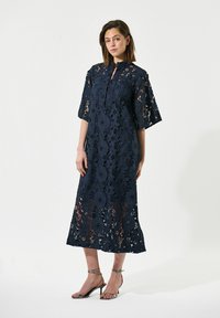 Vestido de encaje azul marino con un cuello alto y mangas anchas, con patrones florales y una longitud por debajo de la rodilla. Combinándolo con tacones metálicos.