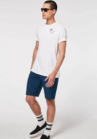 Weißes Baumwoll-T-Shirt mit hochgerollten Ärmeln, marineblauen Shorts, schwarzen Sonnenbrillen und schwarzen Slippern mit weißen Sohlen und gestreiften Socken.