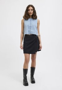 Top din denim fără mâneci, cu tiv desfăcut, asortat cu o fustă neagră înfășurată. Modelul poartă cizme negre până la genunchi. Design simplu, modern.