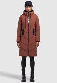 Brun polstret lang frakke med chevronmønster, hætte, frontlynlås, sorte detaljer og to sidelommer. Båret over tætsiddende sorte bukser og støvler.